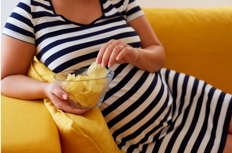 A pregnant woman is laying down on the sofa eating potato chips.