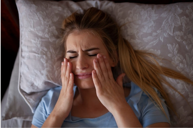 A young woman who's suffering from TMJ (Temporomandibular joint) pain is clenching her jaw in pain in her bed.