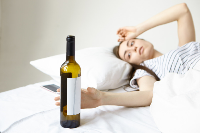 A girl is resting in bed because of a recent feeling of a blackout but is reaching out to drink from her wine bottle. Drinking alcohol is causing her to have split-second blackouts.