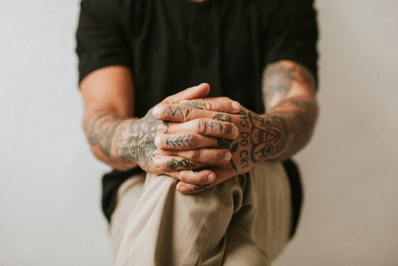 A tattoo artist is sitting down, showing both his hands which are filled with tattoos.