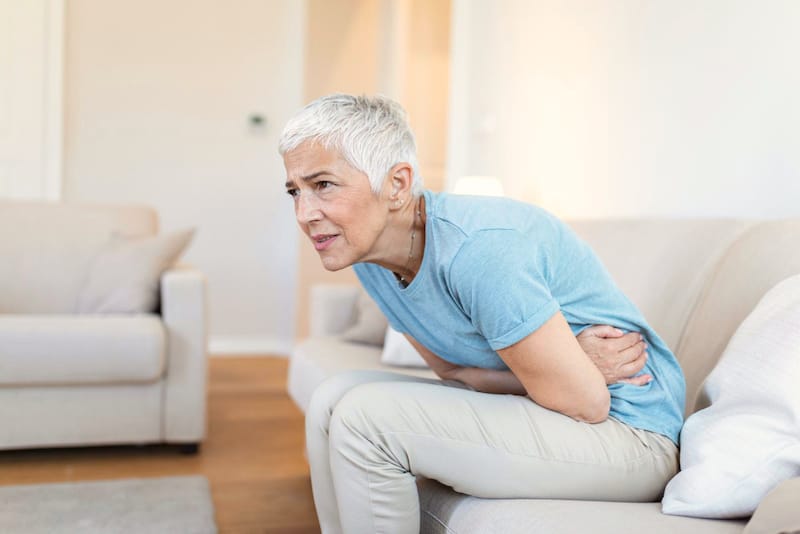 An older woman is sitting on the sofa and clenching her stomach as she's feeling discomfort from her irritable bowel syndrome condition