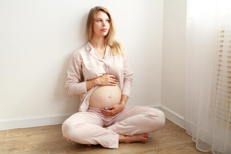 A pregnant woman is sitting down on the floor looking outside on a sunny morning