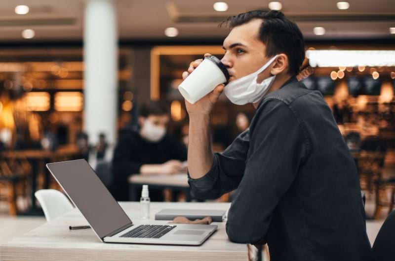 A man with a mask is sitting by the table and sipping coffee while working. 