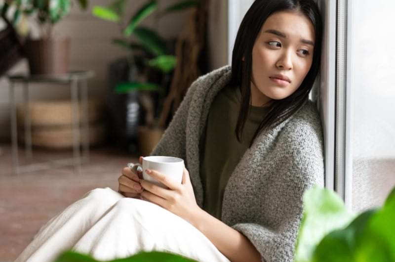 A woman is sitting beside the window deep in thought and drinking coffee.