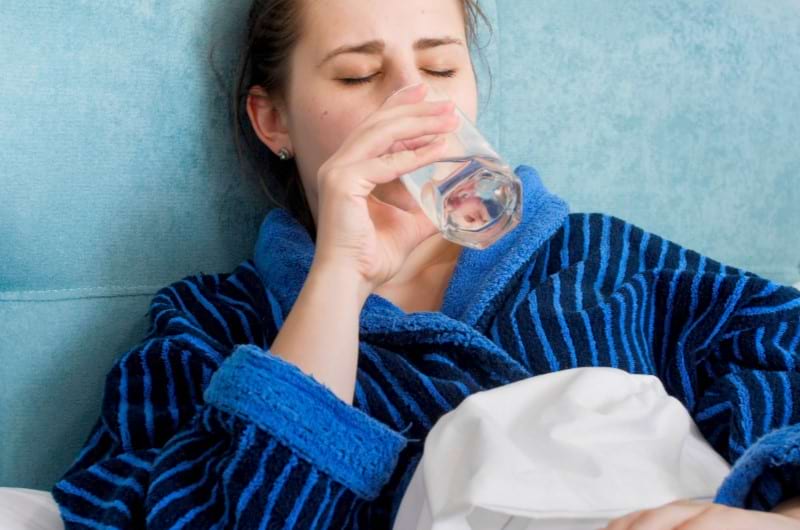 A sick woman is lying in bed and drinking water to keep hydrated.