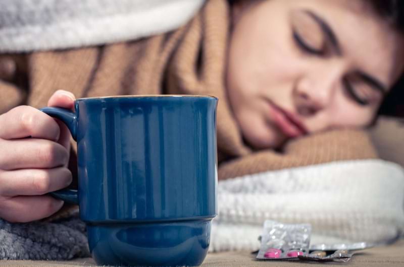 A young woman is lying on the bed sick with a cup of coffee and medicines beside her.