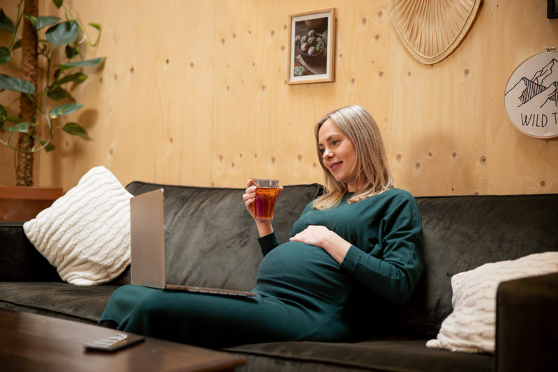 A pregnant woman is sitting down drinking team and watching something on her laptop