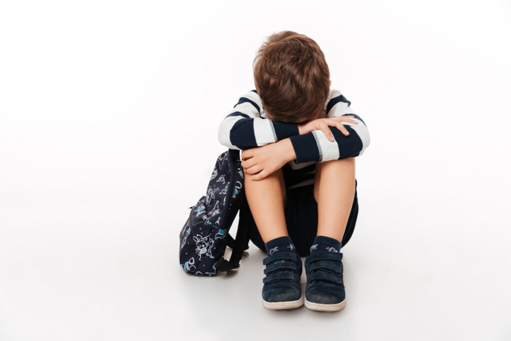 A young boy looks noticeably sad and is sitting down with his head between his legs