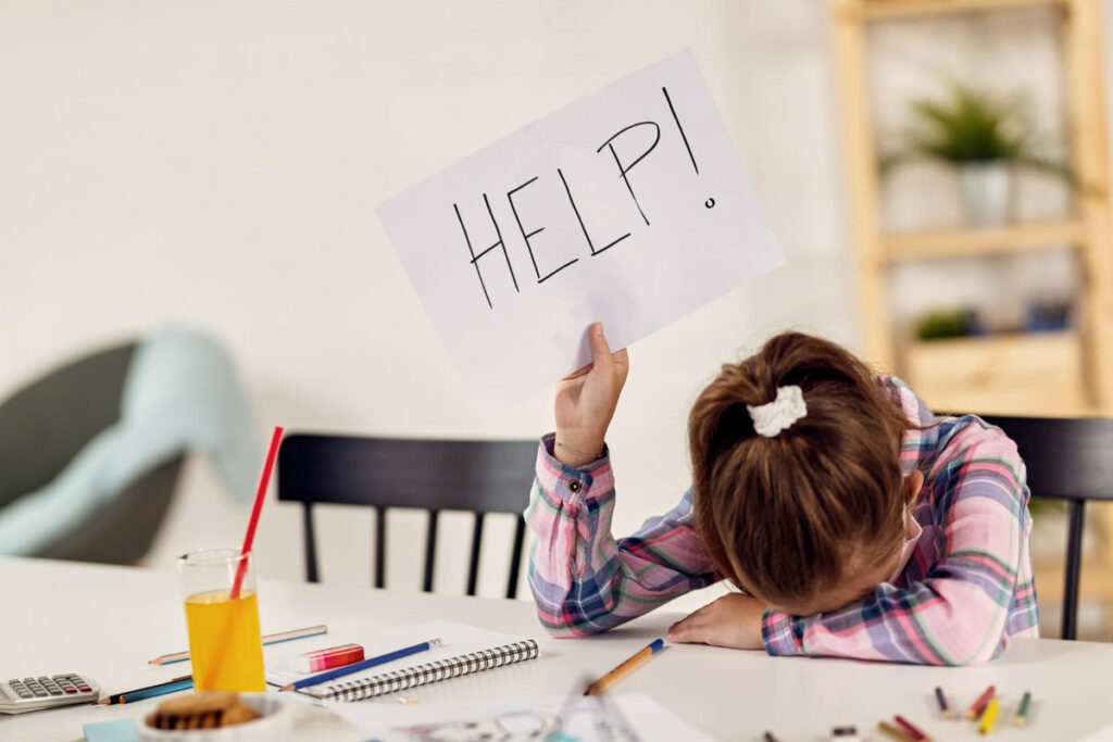 A young girl has her head on her desk with a sign saying "Help" in her hand, she looks defeated